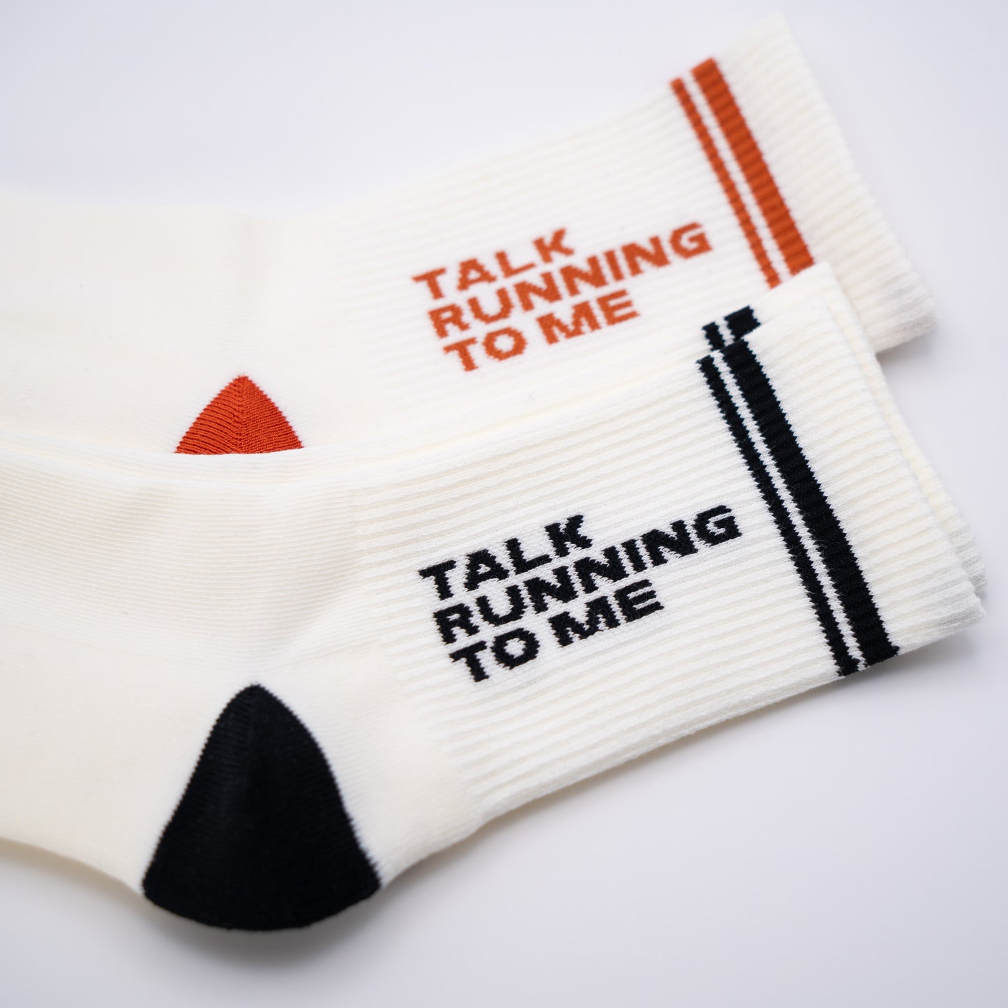 Two white ribbed crew socks with the text 'TALK RUNNING TO ME' printed on them, one in orange and the other in black, shown on a clean white background.