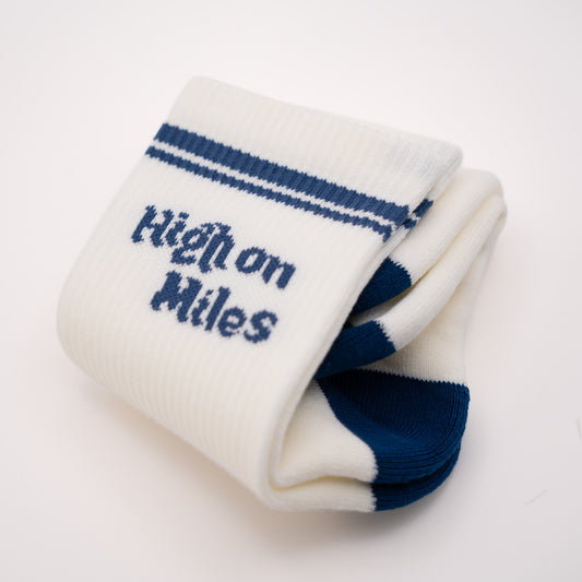 A pair of folded white knit socks with navy blue stripes near the cuff and the words 'High on Miles' knitted in navy on the front, shown on a clean white background.