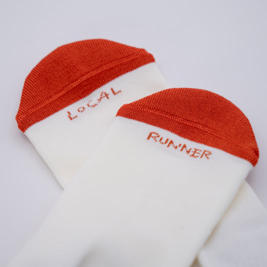 A pair of white athletic socks with rust-orange cuffs and heels, featuring the words 'LOCAL' and 'RUNNER' embroidered in matching rust-orange text on the toes.