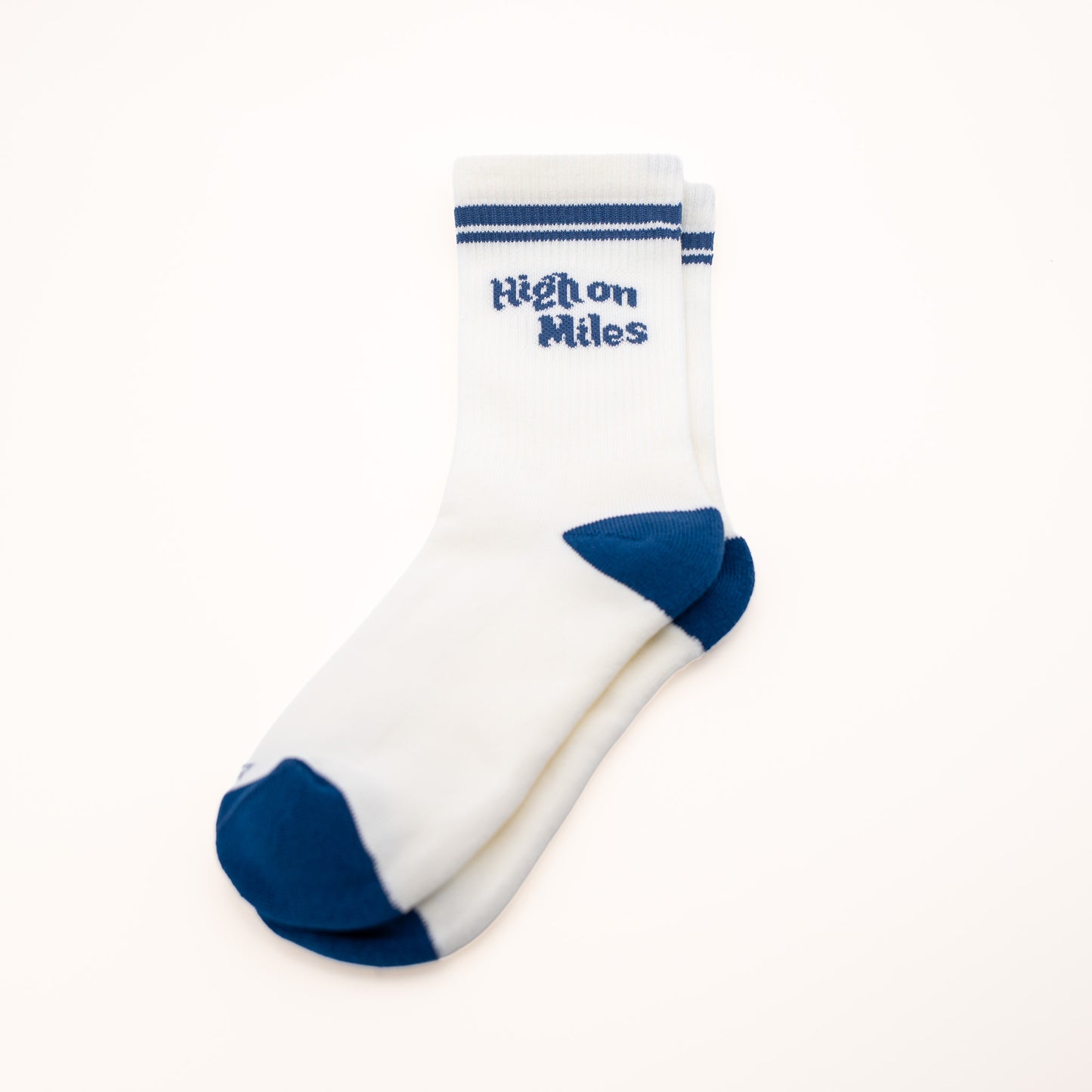 A pair of white crew socks with navy blue accents on the toe, heel, and sole, featuring the text 'HIGH ON MILES' in navy blue on the front cuff.