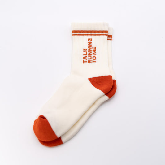 A pair of off-white crew socks with rust-orange accents on the toe, heel, and sole, featuring the text 'TALK RUNNING TO ME' printed vertically on the side. The socks are displayed on a clean, light off-white background.