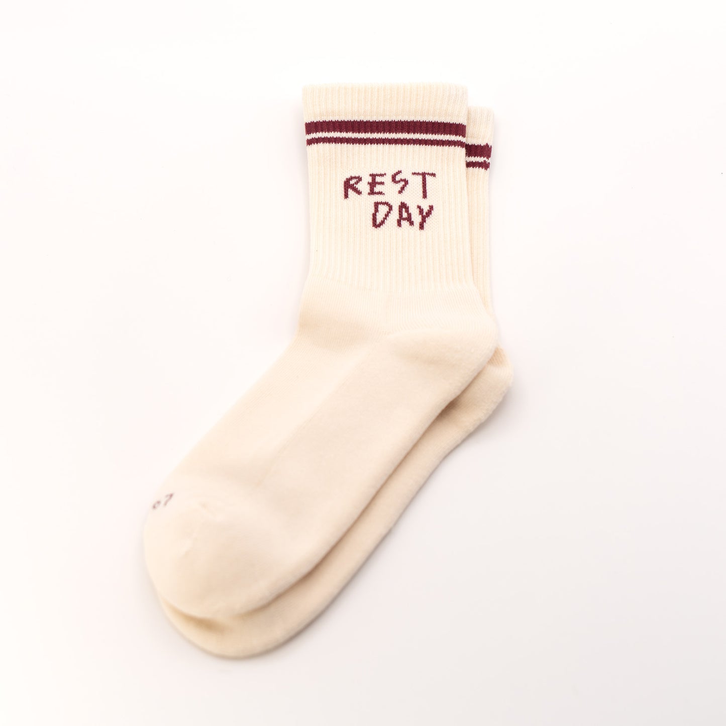 A pair of cream-colored crew socks stacked on a white background, featuring dark red stripes near the ribbed cuff and the words 'REST DAY' printed in dark red on the top sock.