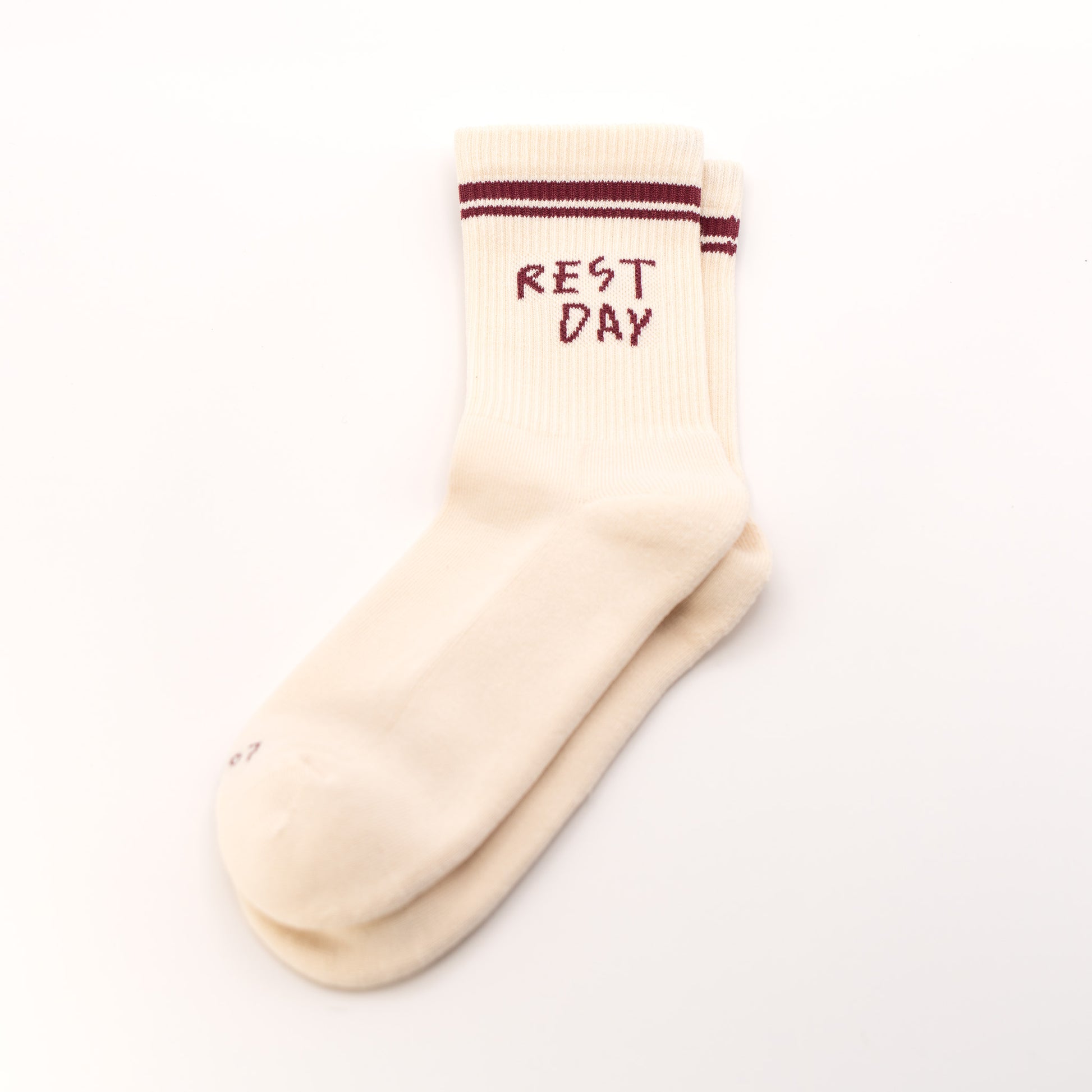 A pair of cream-colored crew socks stacked on a white background, featuring dark red stripes near the ribbed cuff and the words 'REST DAY' printed in dark red on the top sock.