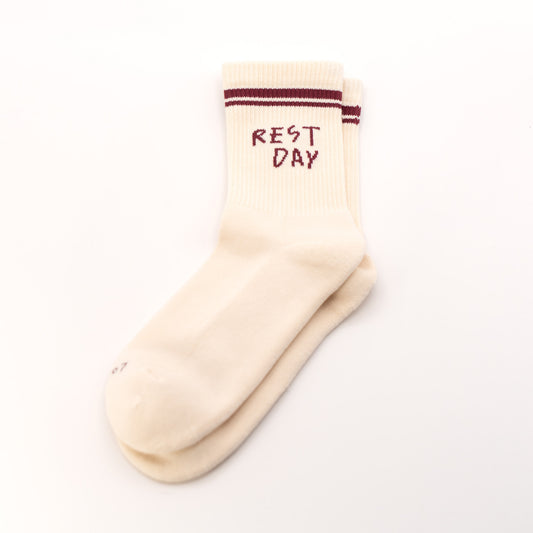 A pair of cream-colored crew socks stacked on a white background, featuring dark red stripes near the ribbed cuff and the words 'REST DAY' printed in dark red on the top sock.