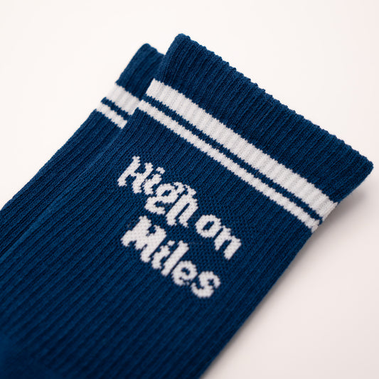 A close-up of a navy blue ribbed sock with white stripes near the cuff and the words 'High on Miles' knitted in white across the front, shown on a plain light background.