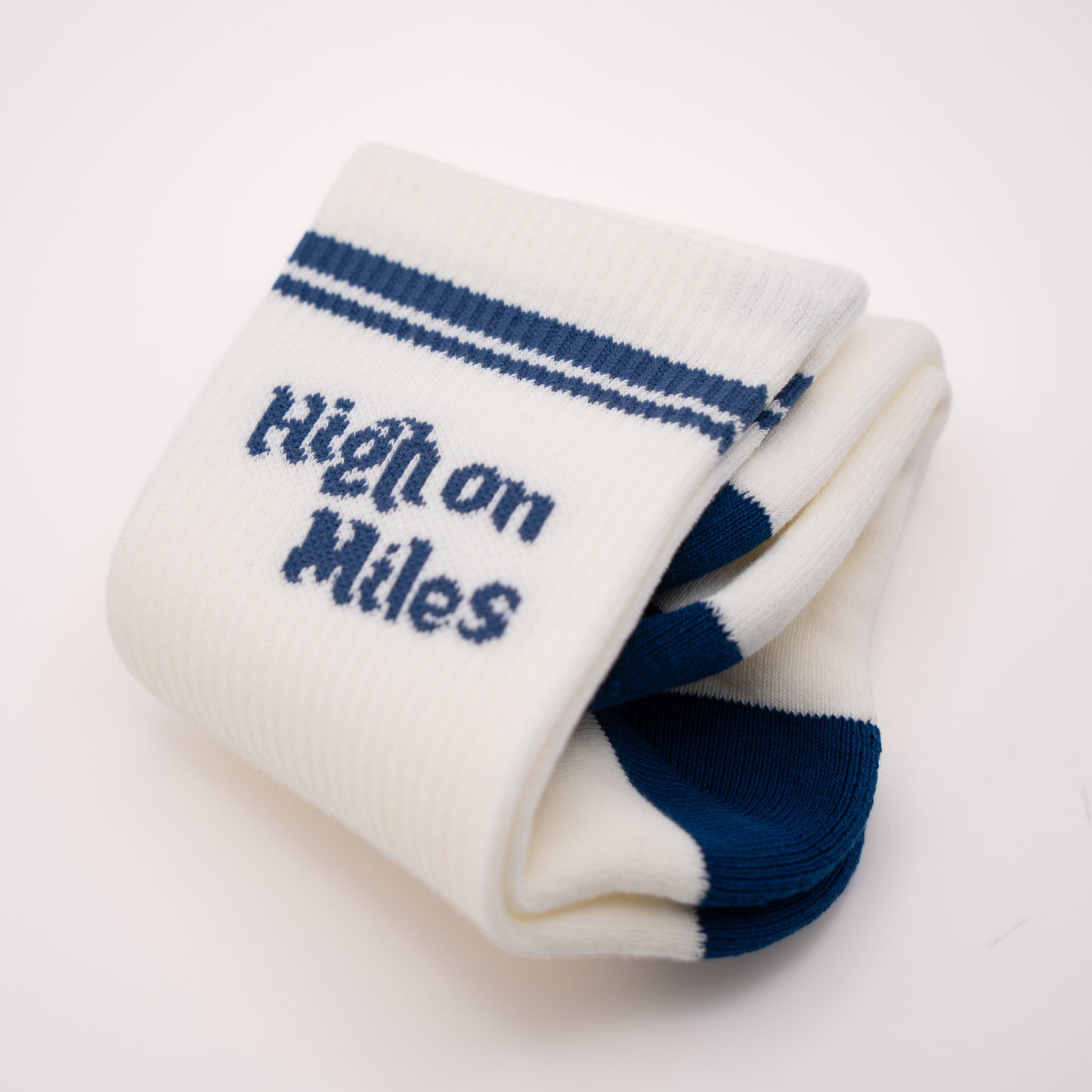 A pair of folded white knit socks with navy blue stripes near the cuff and the words 'High on Miles' knitted in navy on the front, shown on a clean white background.