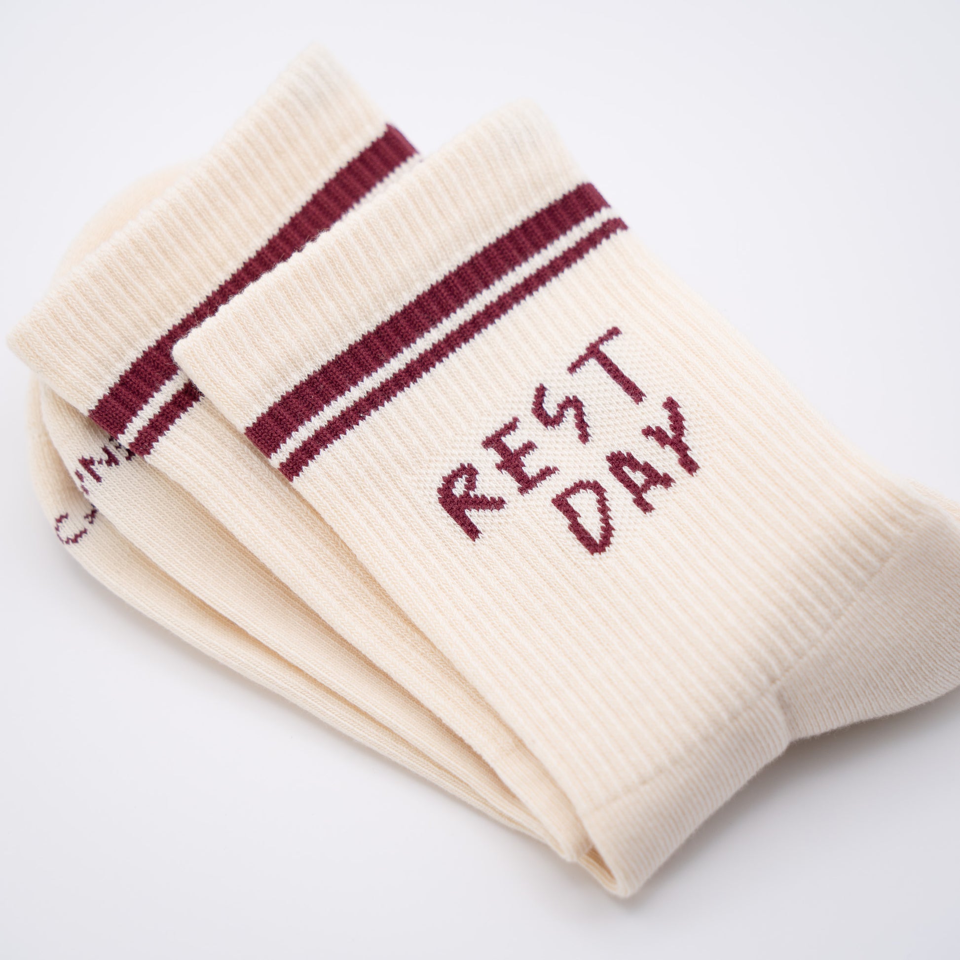A pair of cream-colored ribbed crew socks with burgundy stripes near the cuff and the words 'REST DAY' knitted on the side, shown on a clean white background.