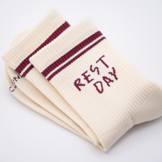 A pair of cream-colored ribbed crew socks with burgundy stripes near the cuff and the words 'REST DAY' knitted on the side, shown on a clean white background.