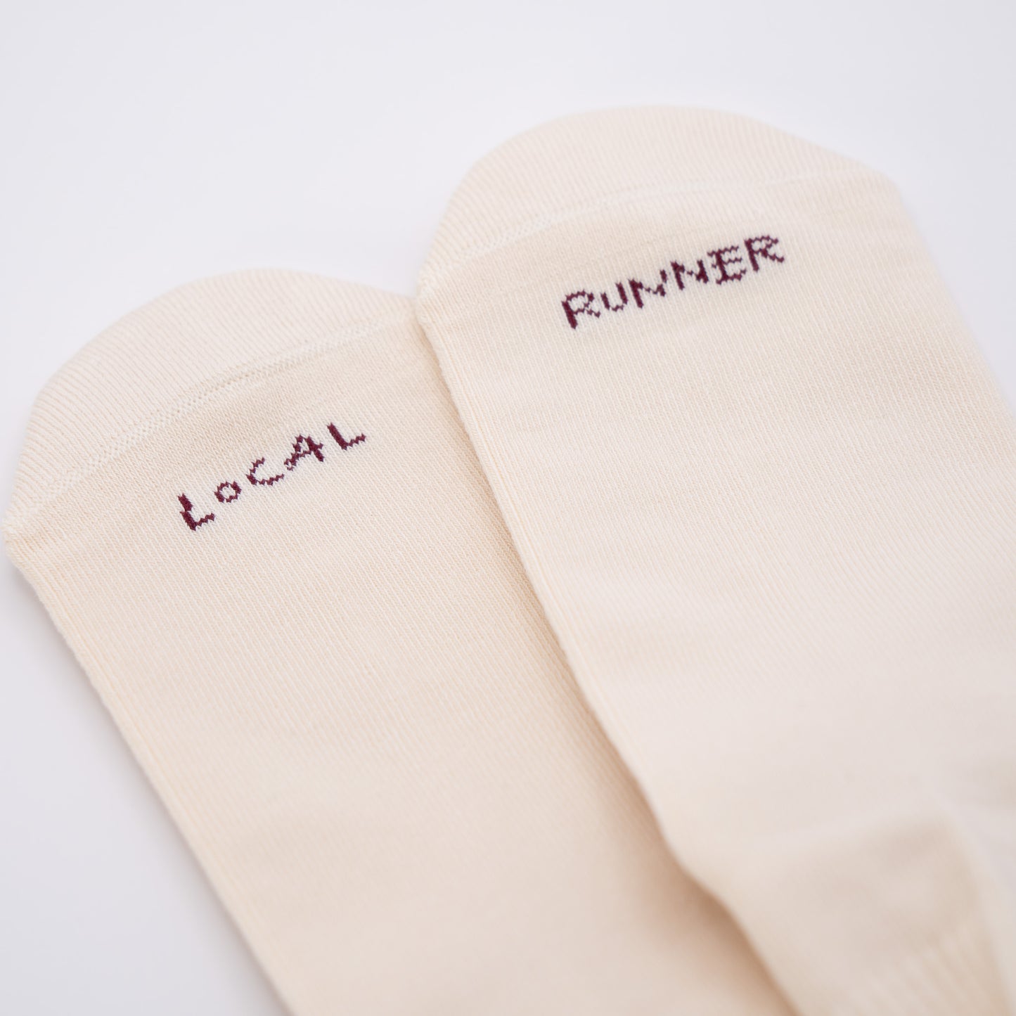 A pair of off-white crew socks with dark red embroidered text reading 'LOCAL' and 'RUNNER' on the toes, shown on a plain white background.