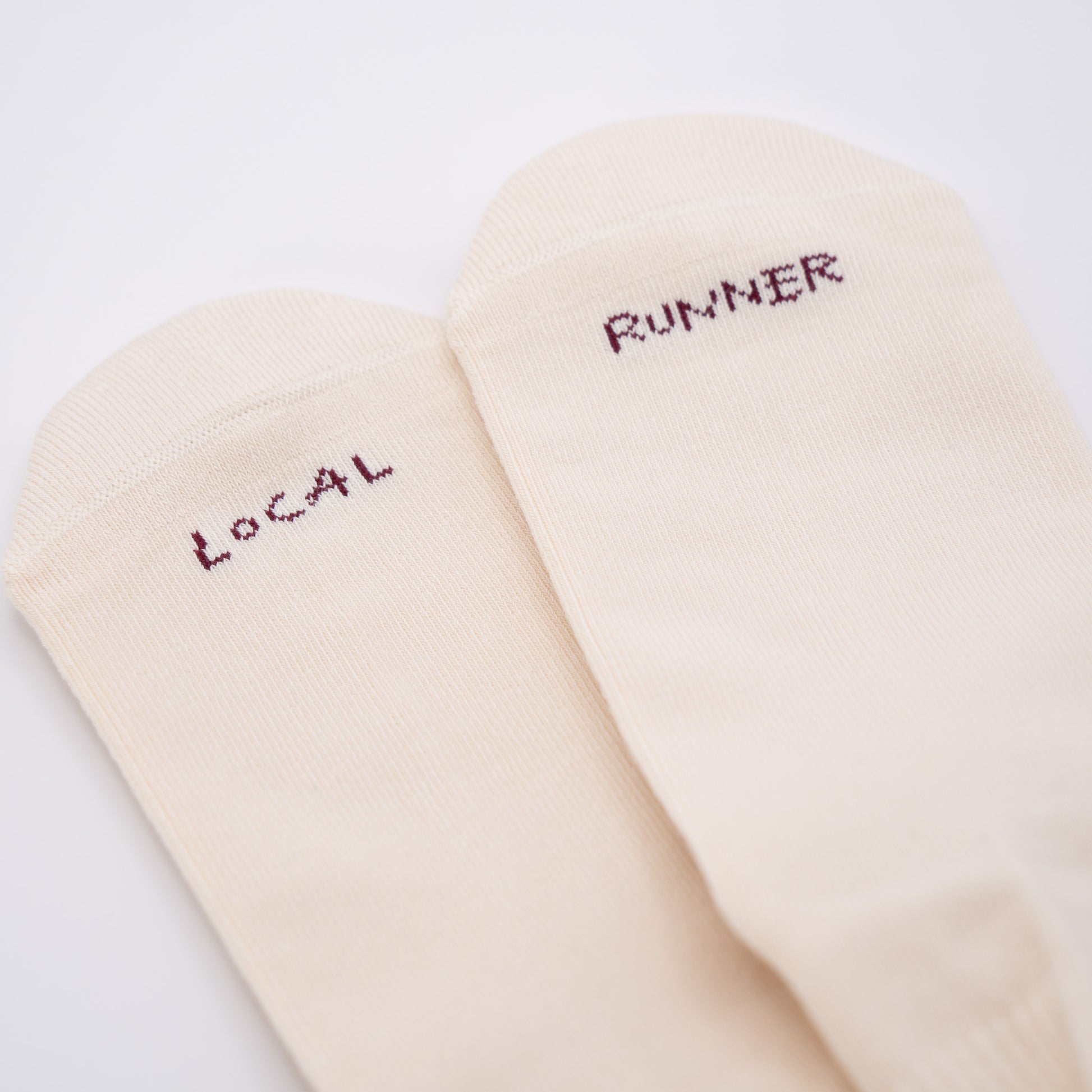 A pair of off-white crew socks with dark red embroidered text reading 'LOCAL' and 'RUNNER' on the toes, shown on a plain white background.