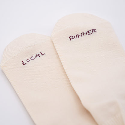 A pair of off-white crew socks with dark red embroidered text reading 'LOCAL' and 'RUNNER' on the toes, shown on a plain white background.