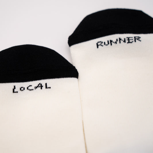 Two off-white crew socks with black toe caps, featuring the words 'LOCAL' and 'RUNNER' printed in black pixel-style text near the cuff, displayed on a clean white background.
