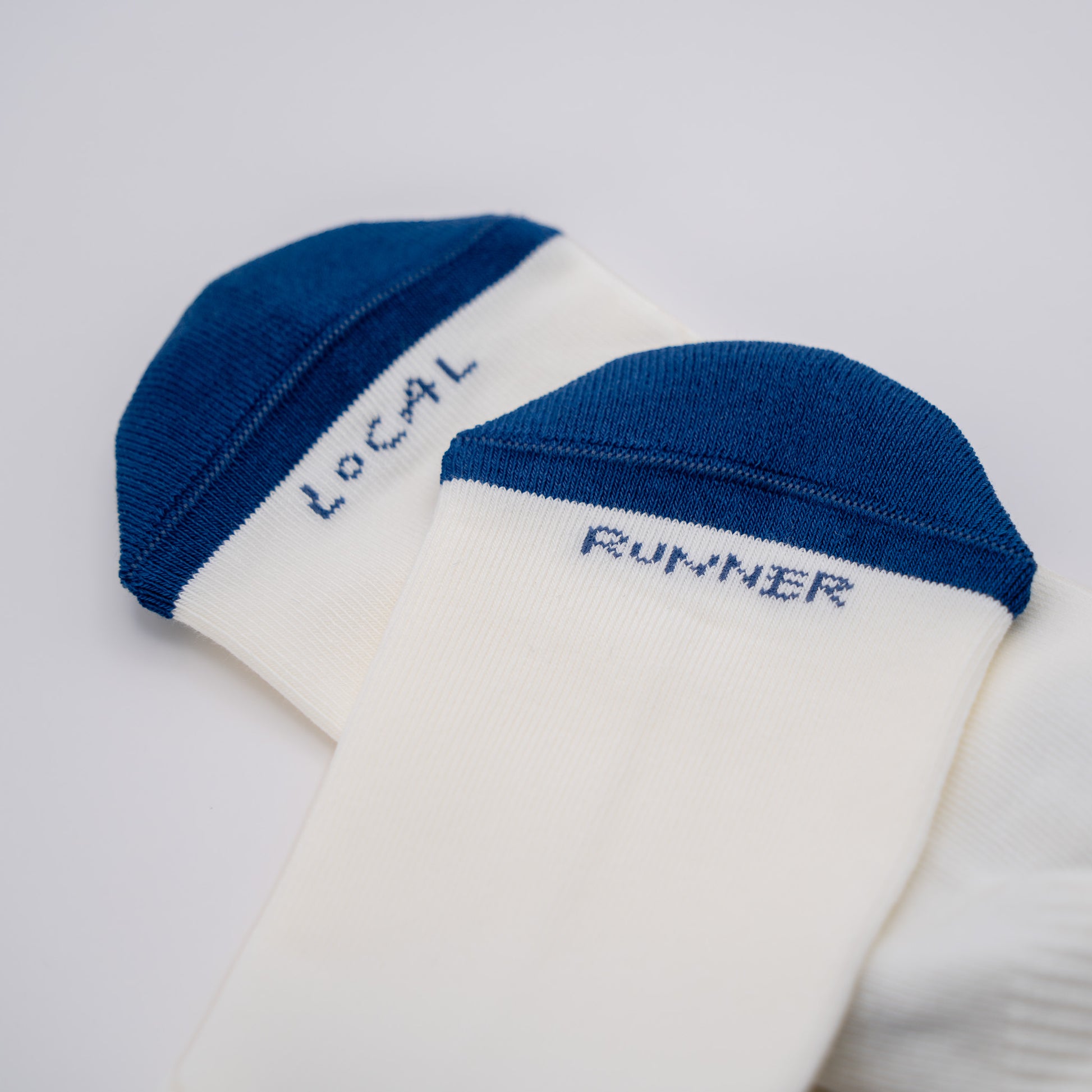 Two white athletic socks with dark blue toe caps, one showing the word 'LOCAL' and the other 'RUNNER' in blue embroidery, on a light gray background.