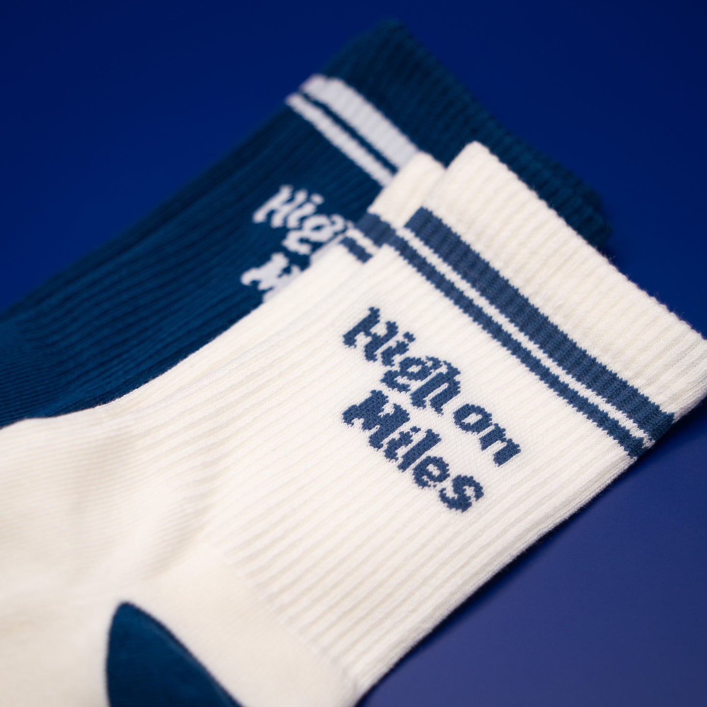 Two ribbed crew socks on a solid blue background, one white with navy blue stripes and 'High on Miles' text, and one dark navy blue with white stripes and the same text.