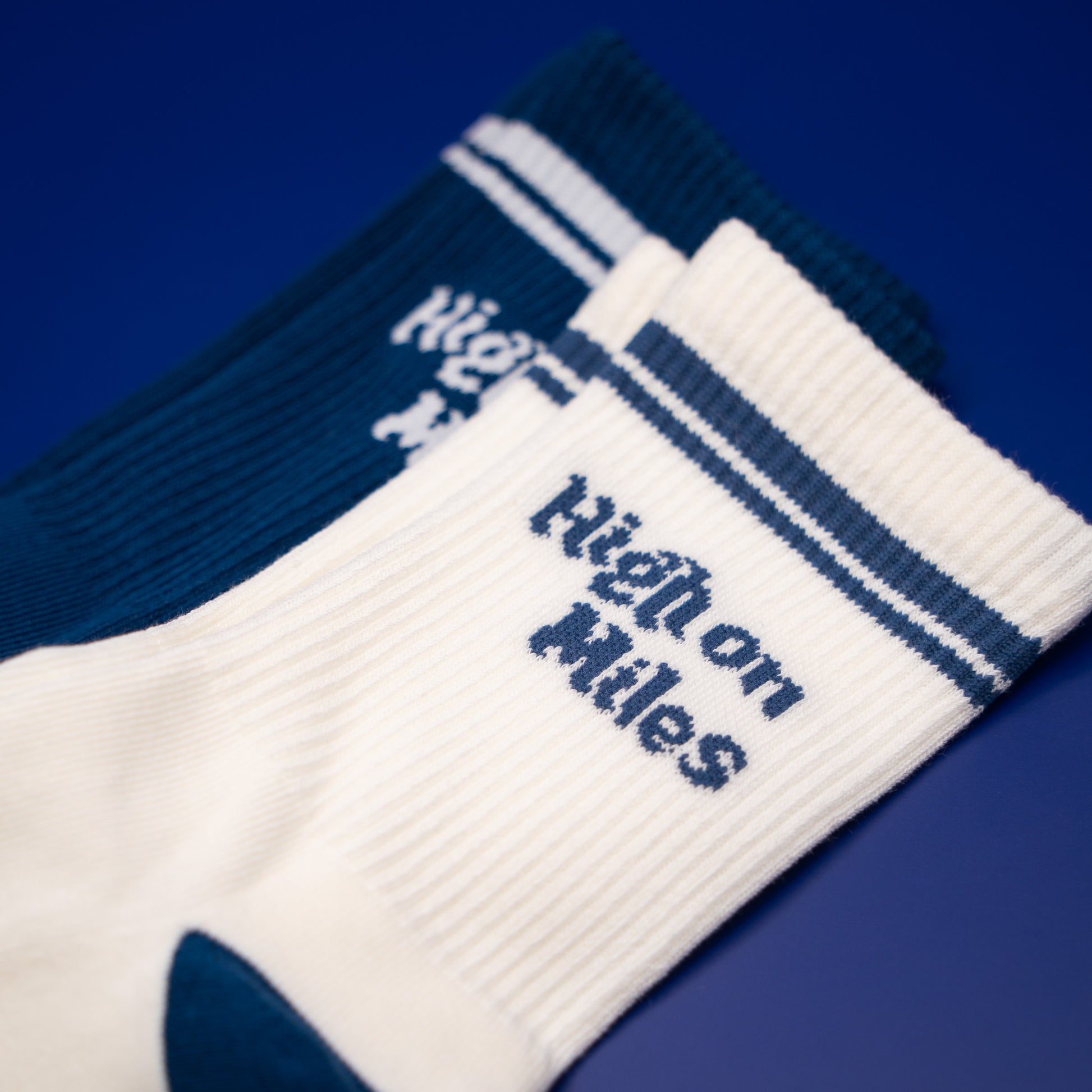 Two ribbed crew socks on a solid blue background, one white with navy blue stripes and 'High on Miles' text, and one dark navy blue with white stripes and the same text.