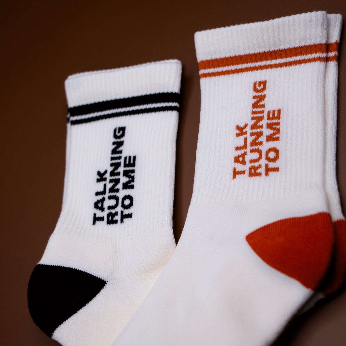 Two white athletic crew socks with 'TALK RUNNING TO ME' printed vertically on the sides, displayed on a dark brown background. One sock has black stripes and black text, while the other has orange stripes and orange text, with orange toe and heel accents.