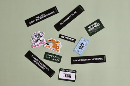 A flat-lay arrangement of various running-themed stickers and name tags on a light sage green background. The collection includes black rectangular name tags with motivational quotes, a light blue ticket stub style tag, a cartoon dog sticker, and a circular logo sticker.