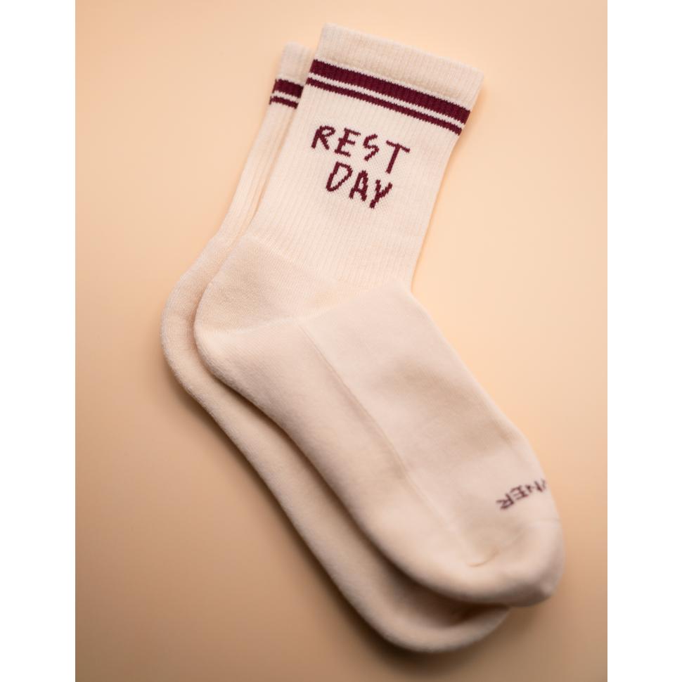 A pair of off-white crew socks with dark burgundy stripes near the cuff and the words 'REST DAY' embroidered in burgundy on the top sock, shown on a warm beige background.