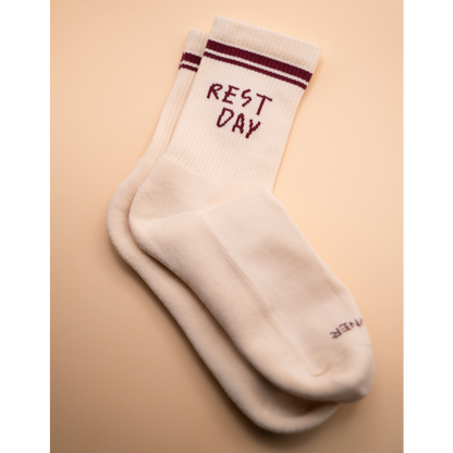 A pair of off-white crew socks with dark burgundy stripes near the cuff and the words 'REST DAY' embroidered in burgundy on the top sock, shown on a warm beige background.